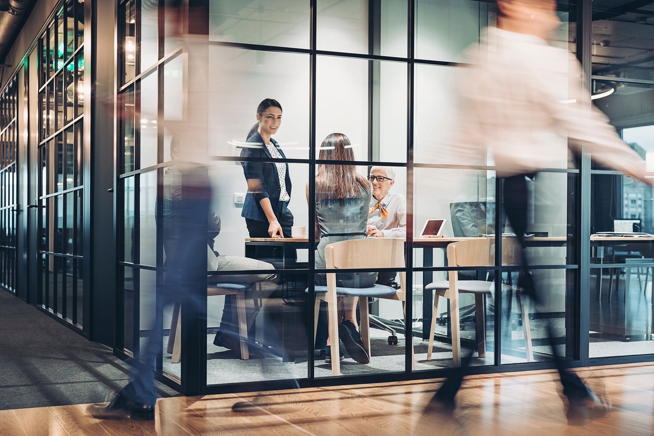 Three women have a discussion in a glass-walled conference room while other workers bustle past in a blur.